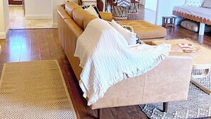 Una sala de estar con un sofá, reposapiés y una mesita de centro. Se observa un comedor con una mesa y sillas al fondo.