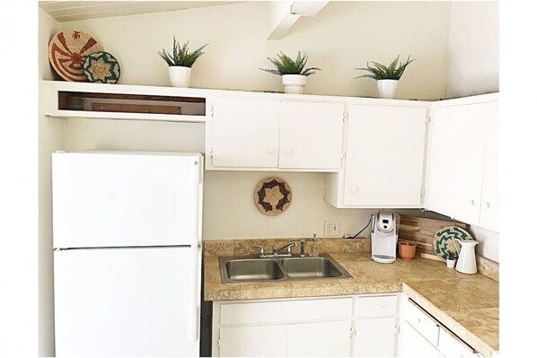 A compact kitchen with white cabinets, a double sink, and a refrigerator. There are potted plants on the countertop and a decorative plate on the wall.