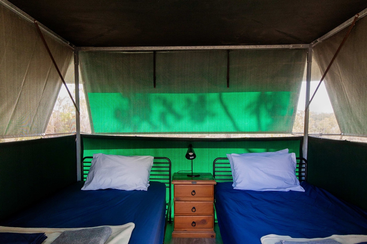 Una carpa con dos camas, cada una con una almohada y una manta. Hay un aparador de madera entre las camas.