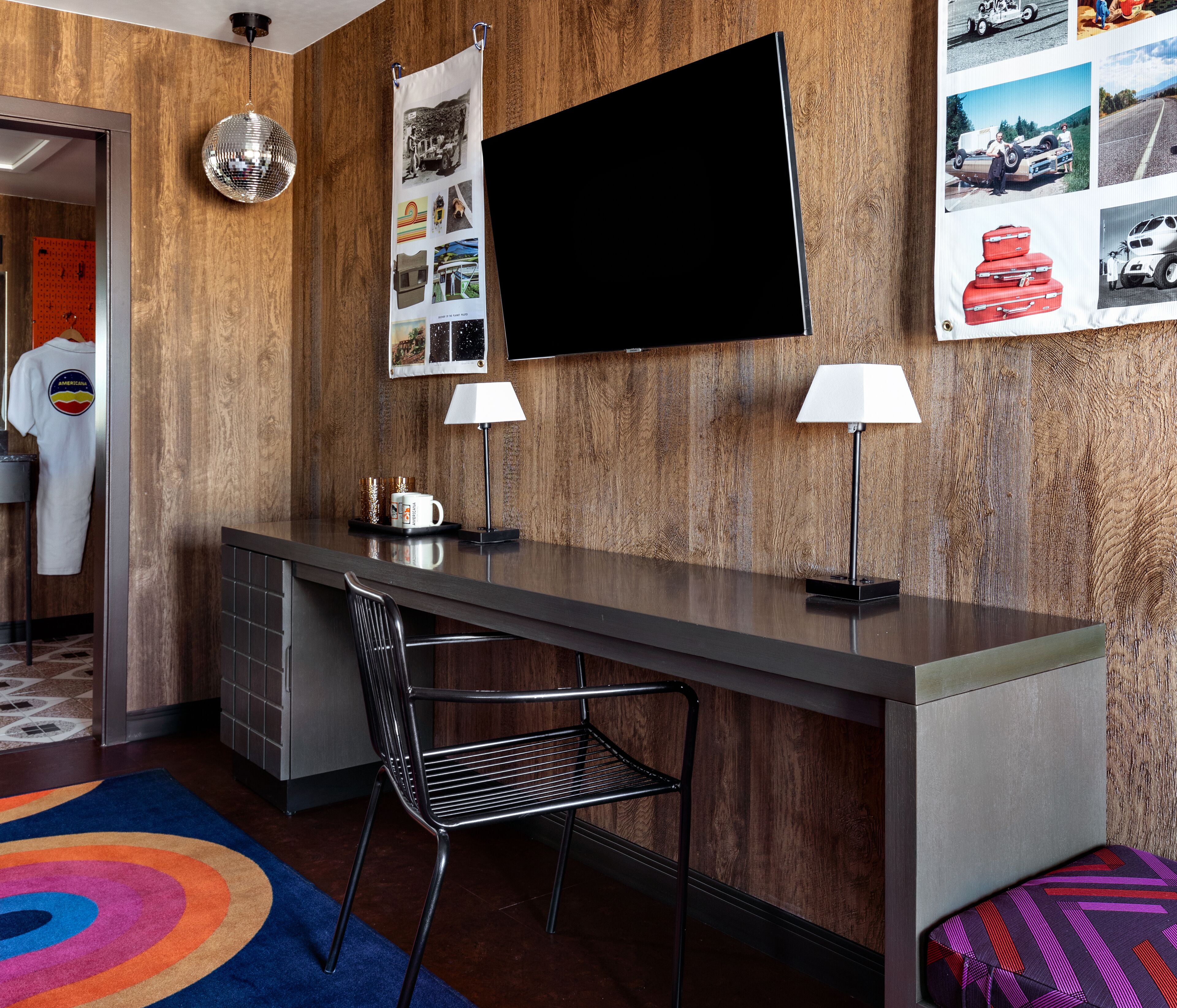 A modern room with a wooden wall, a flat-screen TV, two lamps, a metal chair, and a colorful rug.