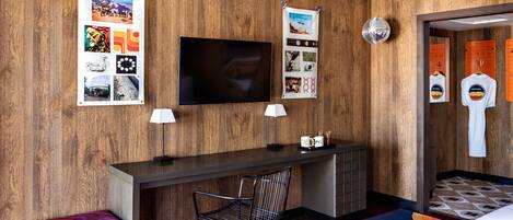 A hotel room with a wooden wall, a flat-screen TV, a desk with a chair, a bench, and a colorful rug.