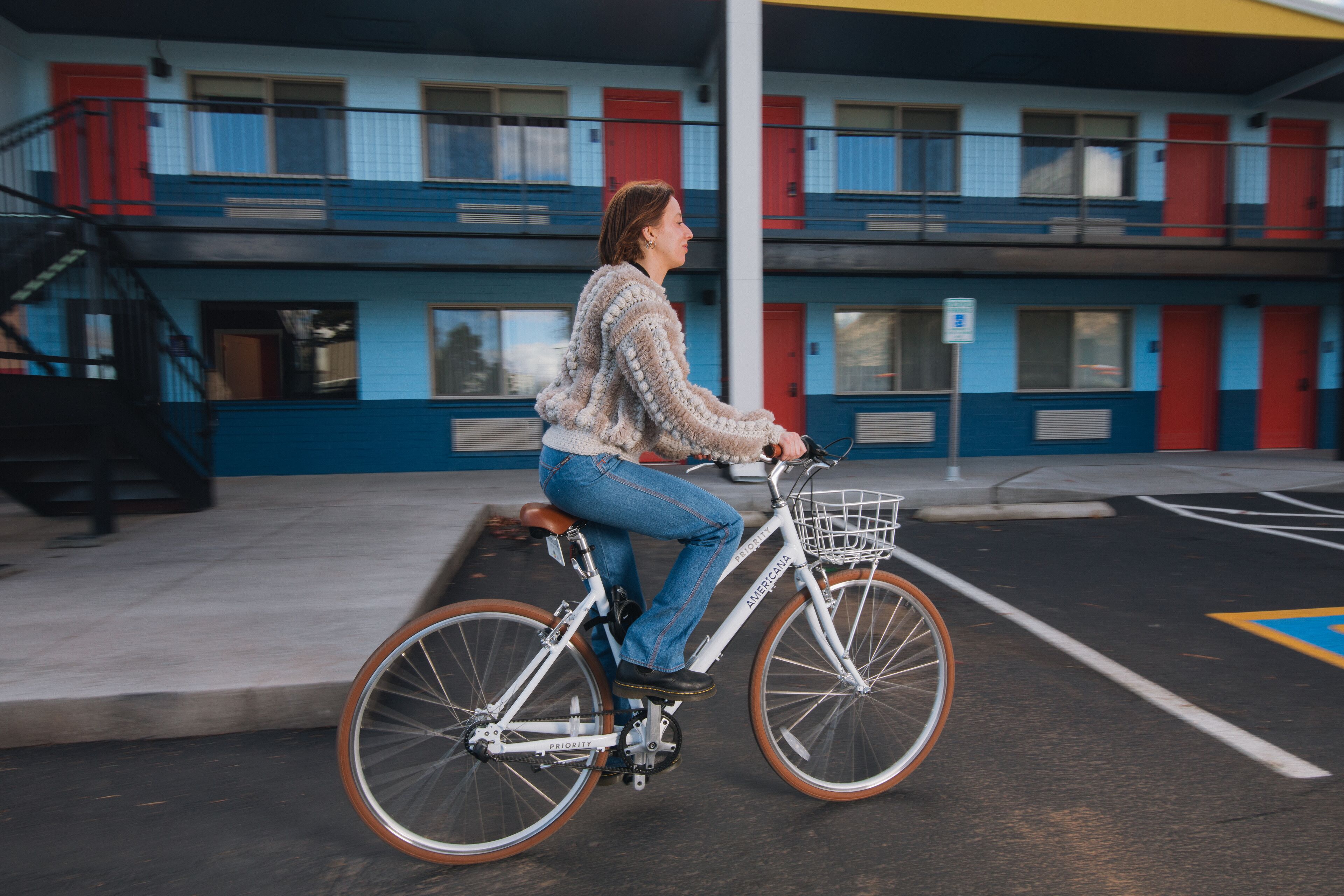 Eine Person fährt mit einem Fahrrad mit Korb durch einen Parkplatz vor einem mehrstöckigen Gebäude mit roten Türen.