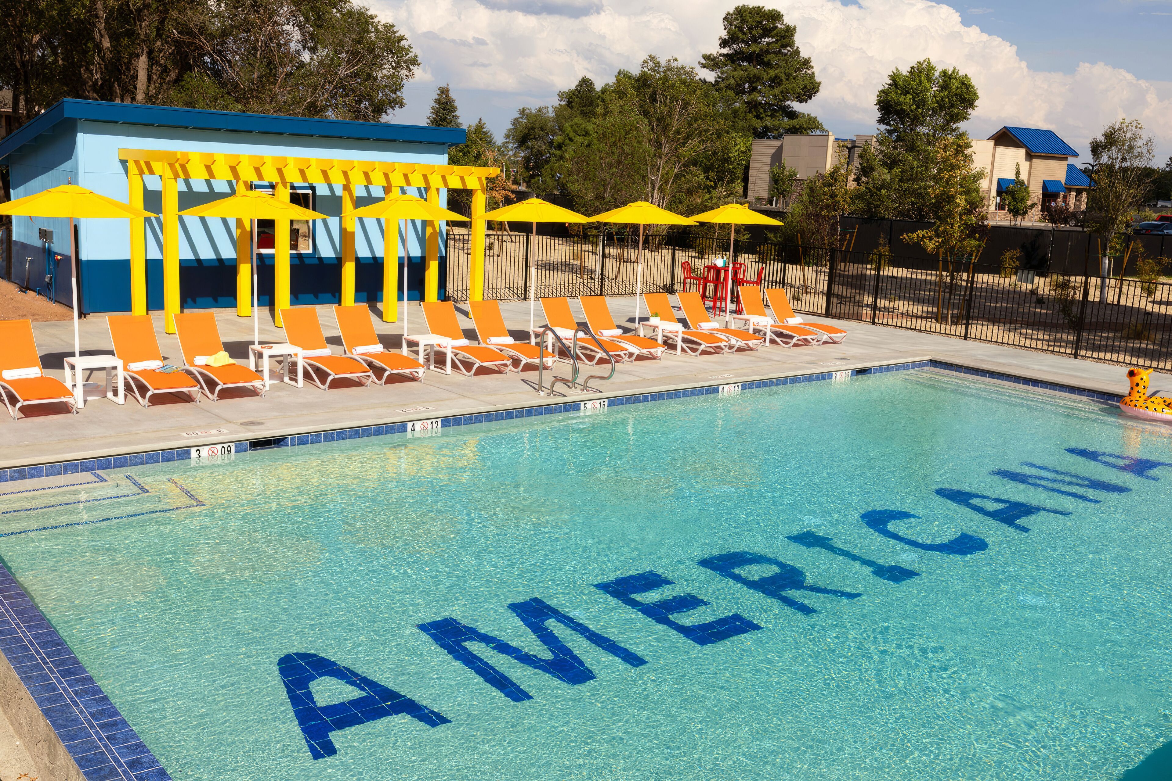 A swimming pool with 'AMERICA' written on the water, surrounded by orange lounge chairs and yellow umbrellas.