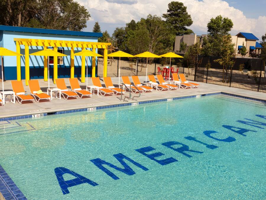 Ein Swimmingpool, dessen Wasseroberfläche das Wort 'AMERICA' zeigt, umgeben von orangefarbenen Liegestühlen und gelben Sonnenschirmen.
