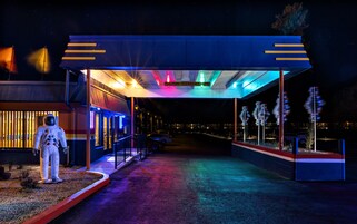 Vista nocturna de la entrada de un edificio con un sendero iluminado con neón, un recinto de cristal y un maniquí de astronauta.