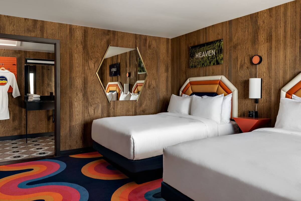 A hotel room with two beds, a mirror, a wall-mounted artwork, and a colorful carpet.