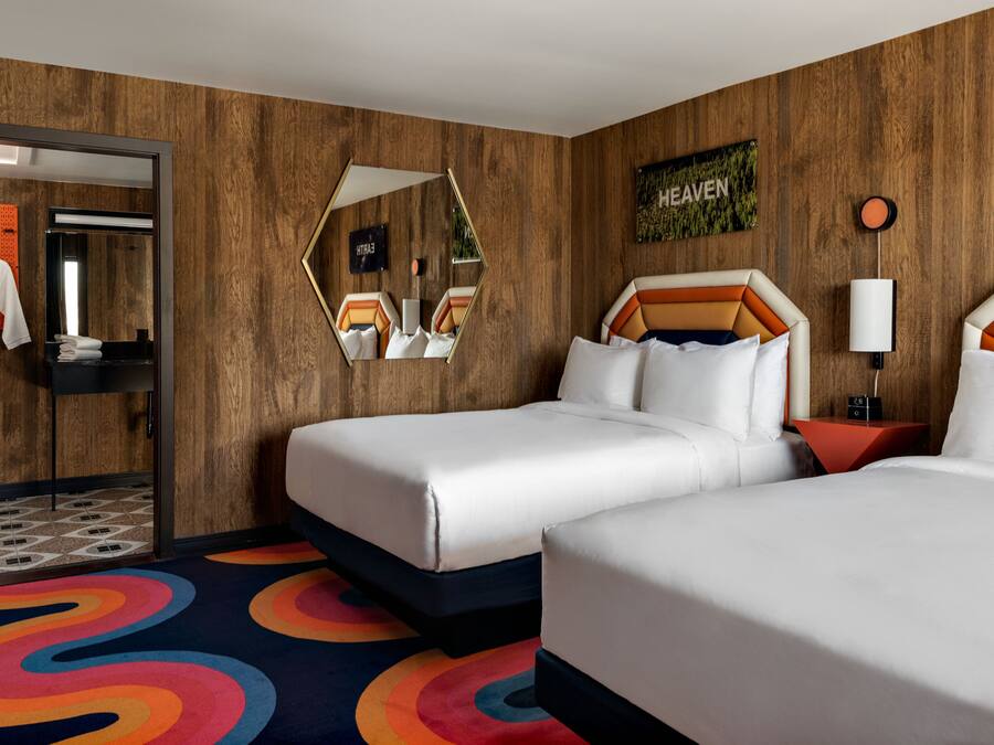 A hotel room with two beds, a mirror, a wall-mounted artwork, and a colorful carpet.