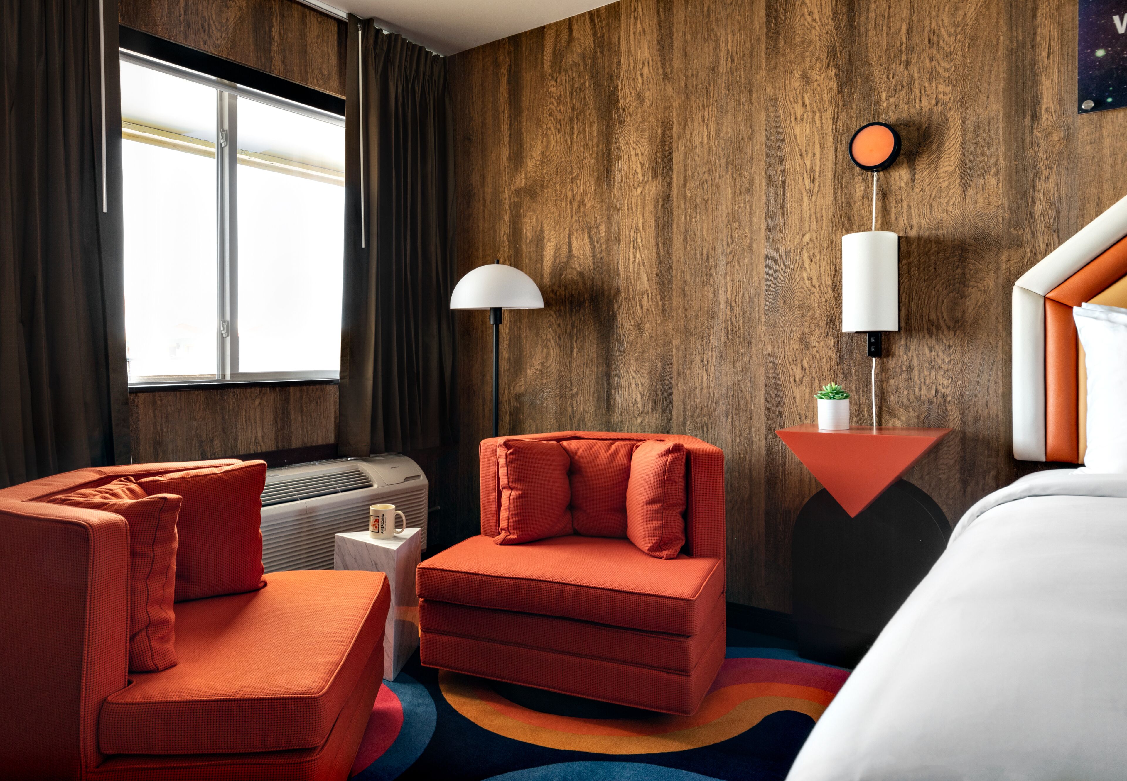 Une chambre d’hôtel avec un mur en bois, un canapé, une chaise, un lit, une table de chevet, une lampe et une fenêtre avec des rideaux.
