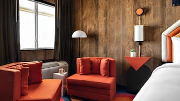 Habitació d'hotel amb paret de fusta, un sofà, una cadira, un llit, una taula de nit, una làmpada i una finestra amb cortines.