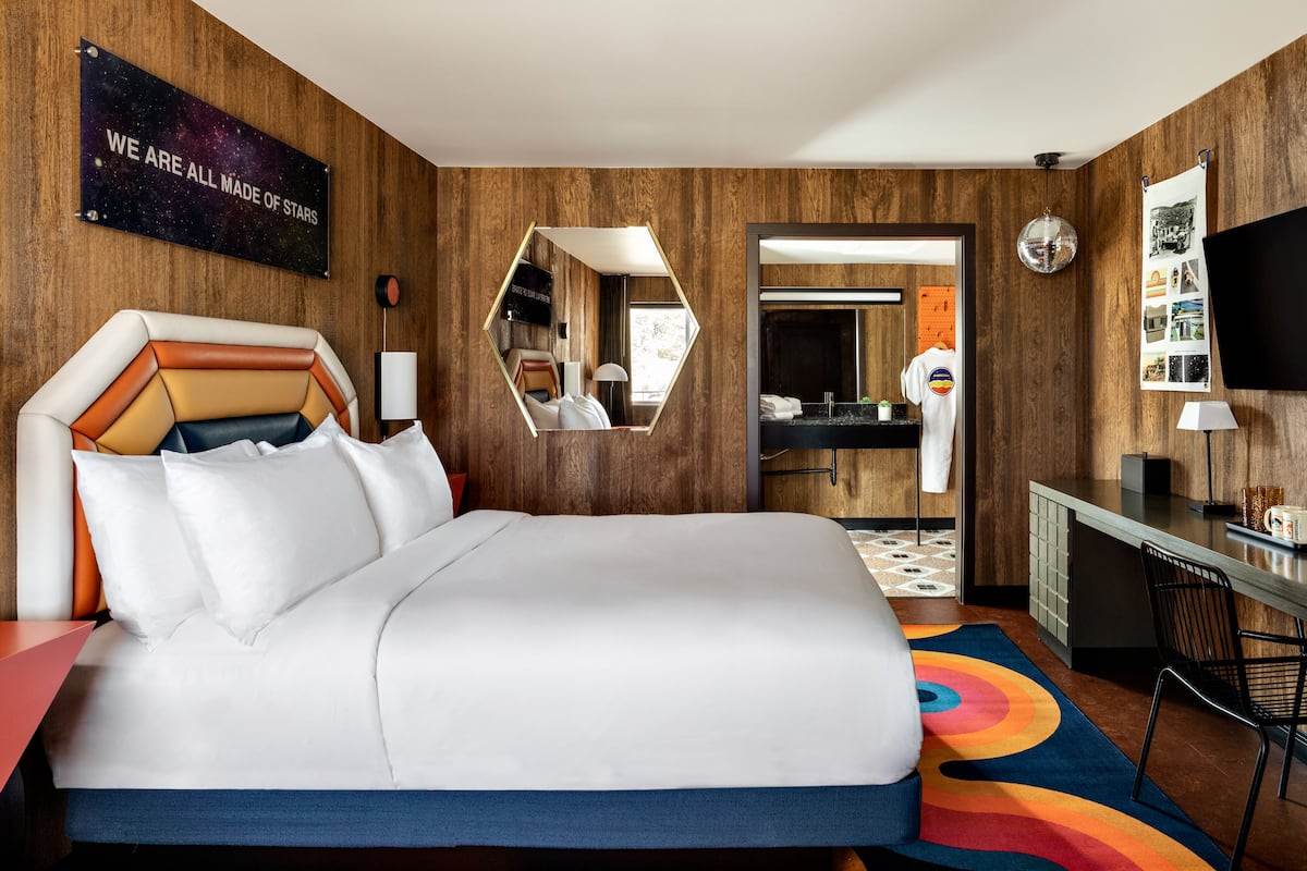 A hotel room with a large bed, a mirror, a desk, a TV, and a bathroom visible through an open door.
