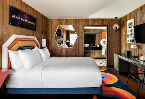 Une chambre d’hôtel avec un grand lit, un miroir, un bureau, une télévision et une salle de bain visible à travers une porte ouverte.