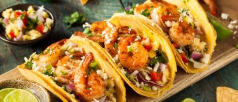 Três tacos de camarão em uma tábua de madeira, com gomos de limão e uma pequena tigela de molho.