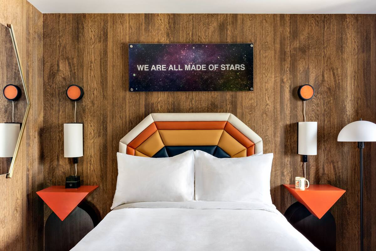 A hotel room with a wooden wall, a galaxy-themed sign, a bed with white linens, and two nightstands with lamps and a coffee cup.