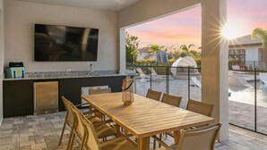 Una cocina moderna con una televisión de pantalla plana, un comedor con mesa y sillas de madera, y vista a una piscina al atardecer.