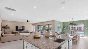 Una cocina moderna con una isla central, un comedor con una mesa larga y una sala de estar con sofá y televisor.