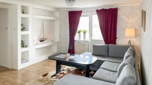 Ein modernes Wohnzimmer mit einem grauen Sofa, einem schwarzen Couchtisch und einem Kuhfellteppich. Zwei rote Vorhänge, eine Pendelleuchte und eine Wandlampe sind vorhanden.