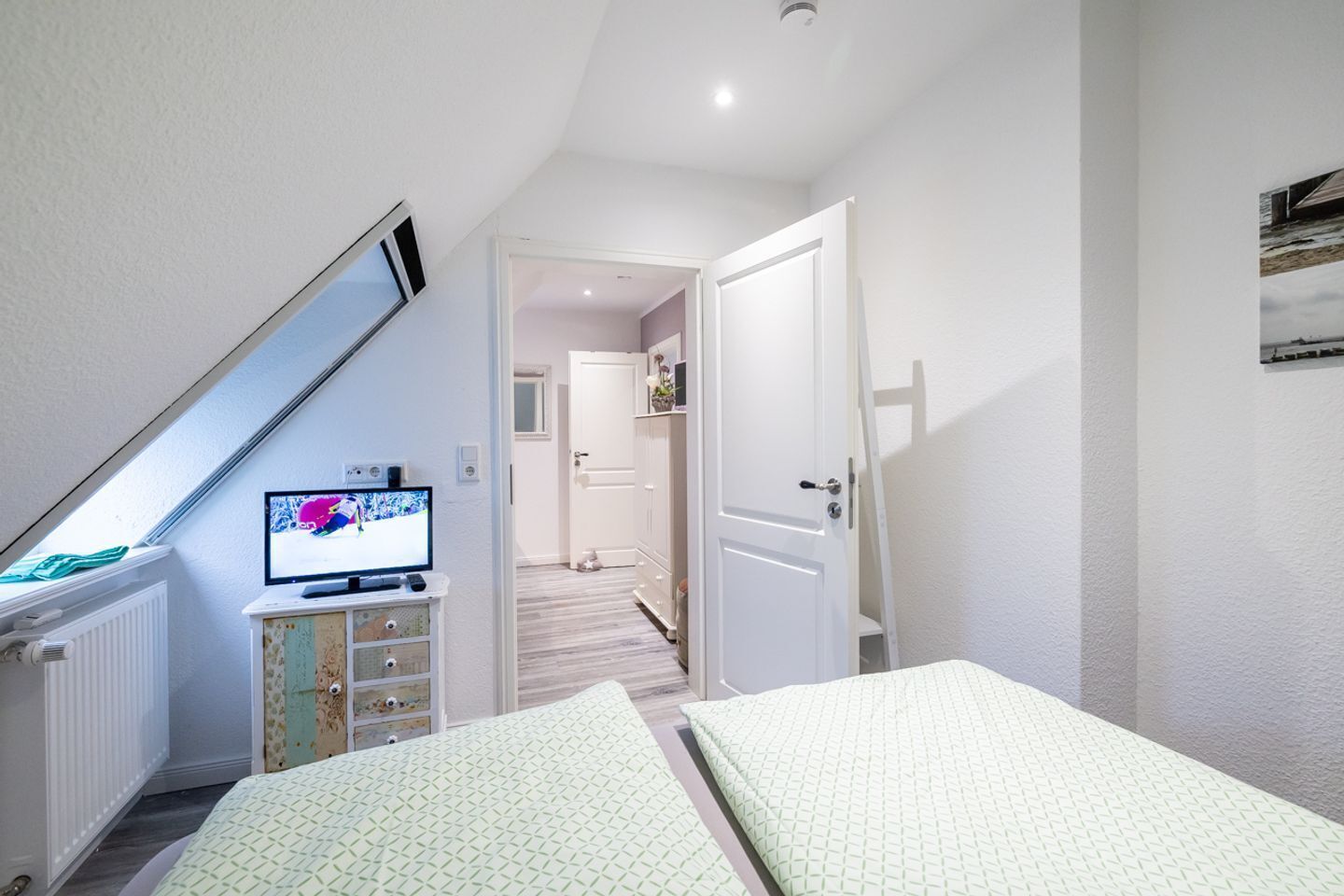 A bedroom with a bed, a television on a dresser, a door leading to another room, and a window with a view of a building.