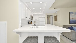 Una cocina moderna con armarios blancos, una isla grande con taburetes y vistas al área de estar con un sofá y obras de arte.