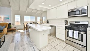 A modern kitchen with white cabinetry, a central island, a built-in oven, and a view of the beach through large windows. - Beachfront home with Sweeping Ocean Views, AC, Walk to the Pier, Restaurants, Shops, Activities (Newport Beach)