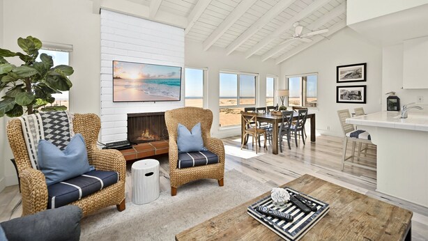A spacious living room with wicker furniture, a fireplace, and a large TV displaying a beach scene. - Beachfront home with Sweeping Ocean Views, AC, Walk to the Pier, Restaurants, Shops, Activities (Newport Beach)