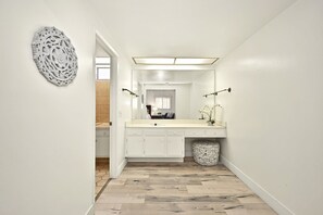 Un couloir étroit mène à une salle de bain équipée d’un meuble-lavabo blanc, d’un miroir et d’un évier. Un élément décoratif orne le mur sur la gauche.