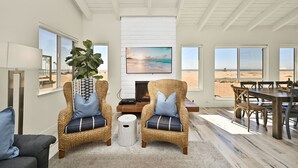A living room with wicker chairs, a fireplace, and large windows overlooking a beach. - Beachfront home with Sweeping Ocean Views, AC, Walk to the Pier, Restaurants, Shops, Activities (Newport Beach)