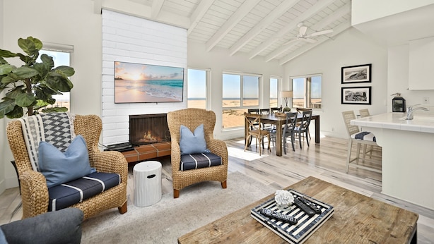 A spacious living room with wicker furniture, a fireplace, and a large TV displaying a beach scene. - Oceanfront Home on the Boardwalk w/ 3 car Garage, AC, Giant Patio, Ocean Views, Walk to Pier (Newport Beach)