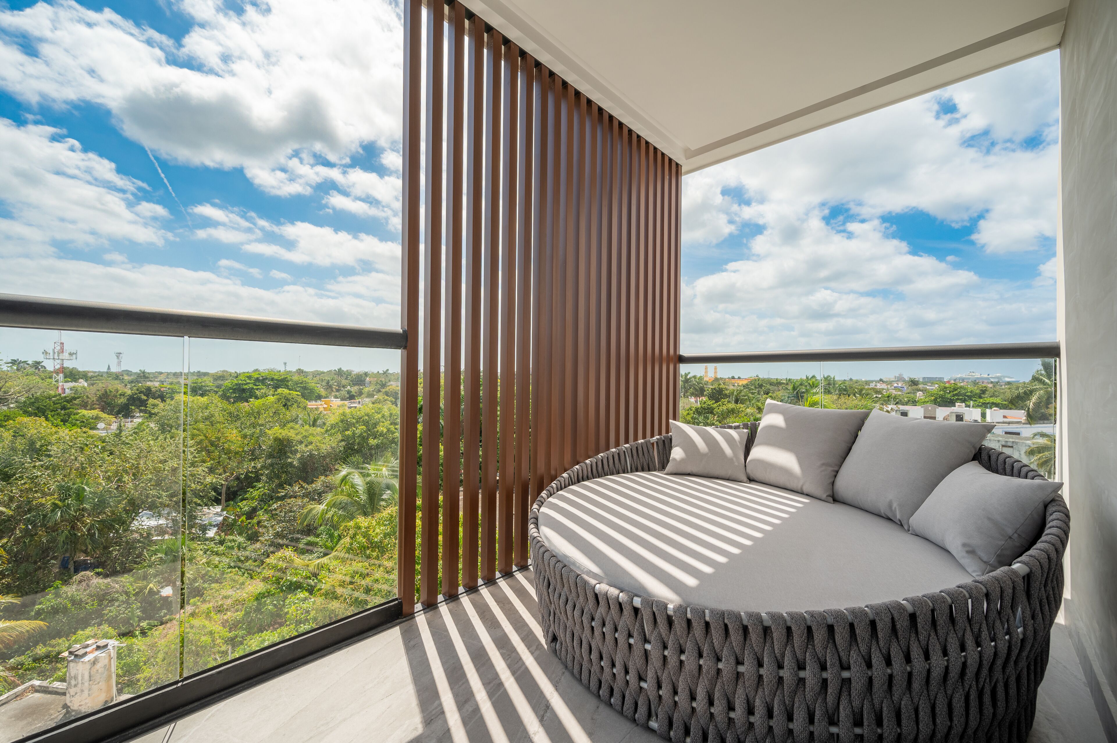 Un balcón con un sofá redondo de mimbre, un panel divisor de listones de madera y vistas a un paisaje verde con edificios a lo lejos.