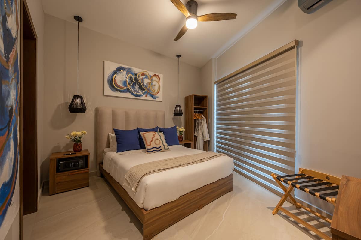Une chambre à coucher avec un lit, un ventilateur de plafond, un tableau, une table de chevet et un banc en bois.