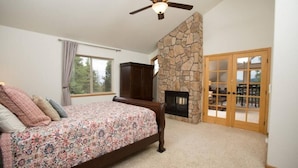 A bedroom with a bed, a stone fireplace, a ceiling fan, and a view of the outdoors through a window. - Wrap-Deck Hot Tub w/ Mt. Sherman Views - Fire Pit, Wildlife (Fairplay)