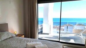 Une chambre à coucher avec un grand lit, une table de chevet avec une lampe et une vue sur la mer par une grande fenêtre.