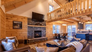 A cozy log cabin interior with a stone fireplace, leather sofas, a dining area, and a kitchen with a large window. - Ursa Minor - Long Range Views, hot tub, Peloton, more at Beautiful 3bd/2ba home on Sugar (Banner Elk)