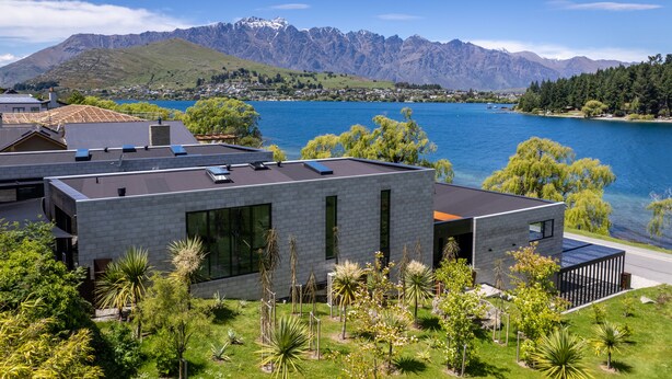 A modern house with a flat roof, large windows, and solar panels, surrounded by lush greenery and a lake with mountains in the background.