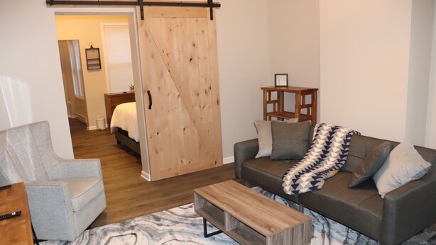 A modern living room with a sliding barn door, a gray armchair, a wooden coffee table, and a gray sofa with patterned throw blankets.