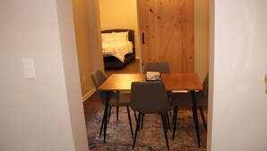 A small dining area with a table and four chairs, a wooden door, and a bed in the background.