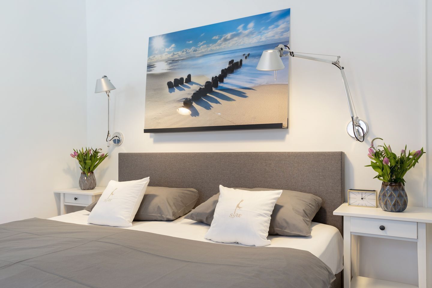 A modern bedroom with a large bed, two bedside tables, a wall-mounted lamp, and a painting of a beach scene.