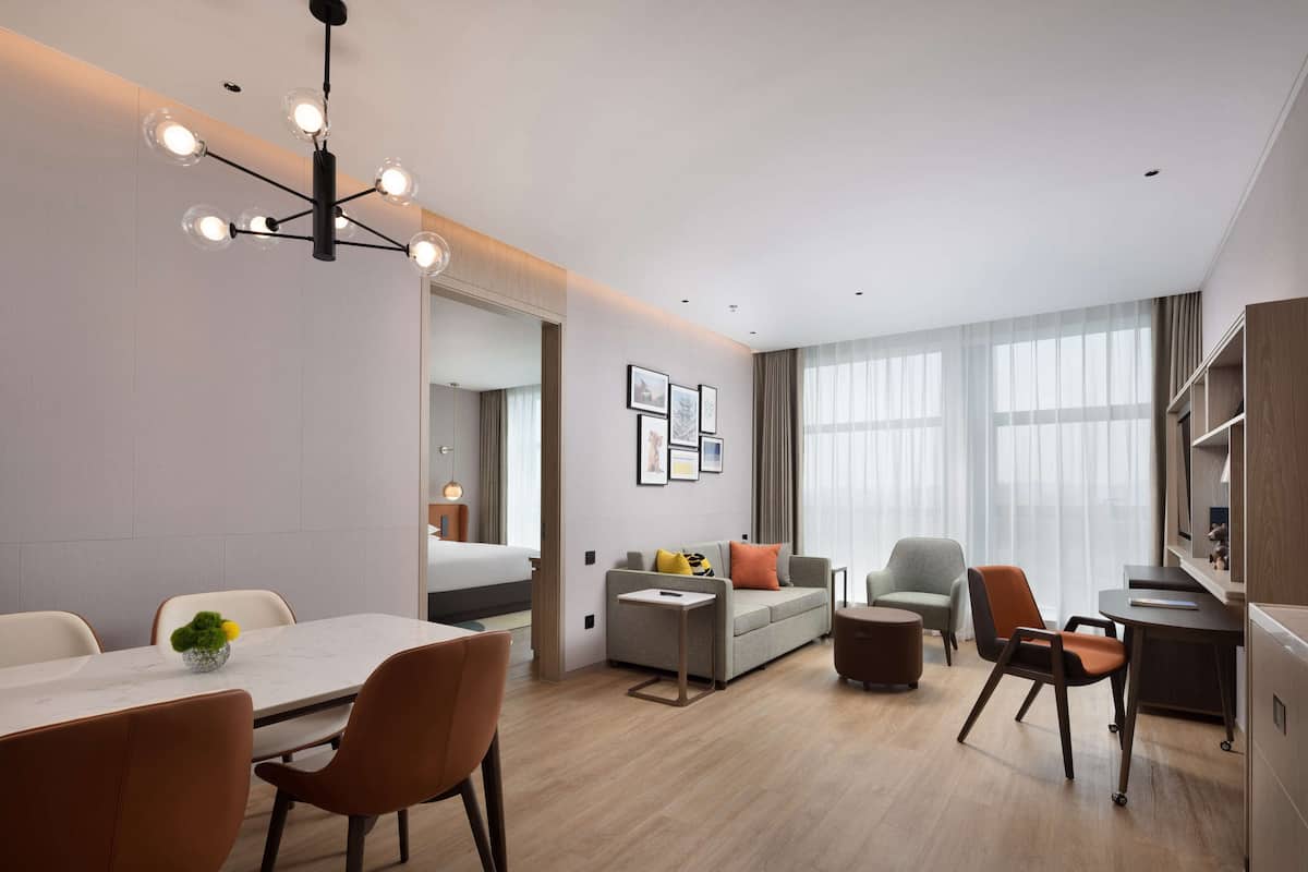 A modern hotel room with a dining area, a sofa, a desk, and a large window.