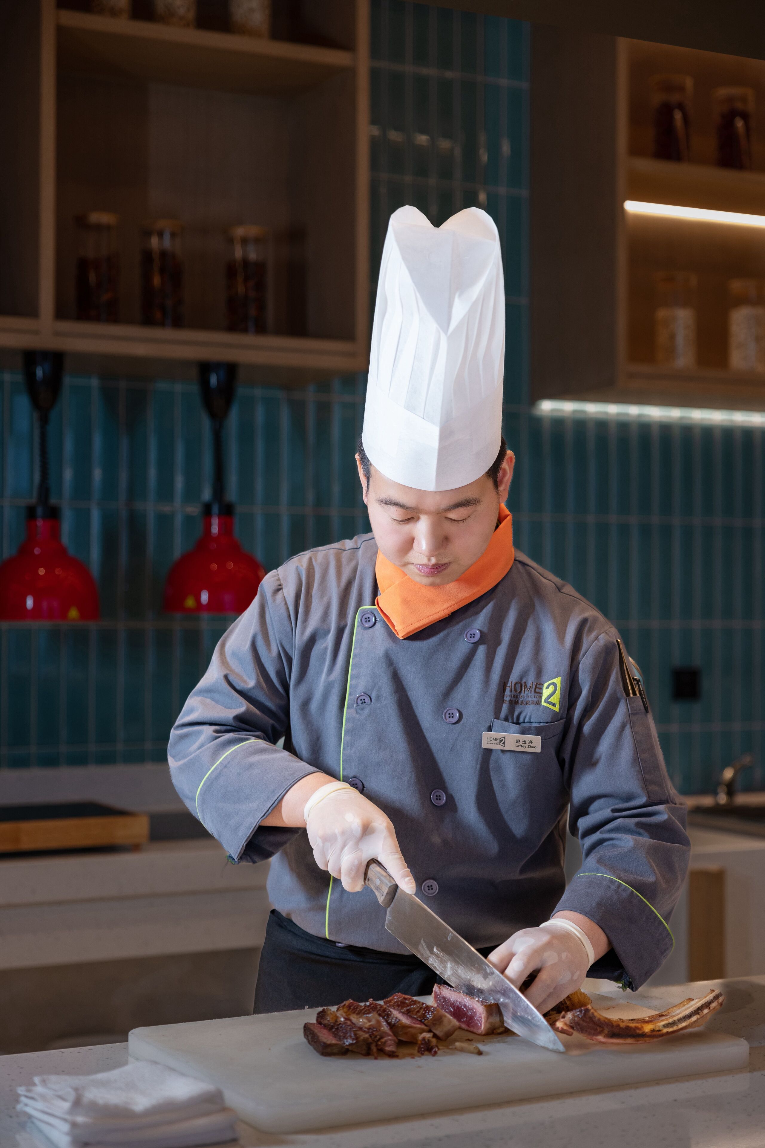 A chef in a uniform cutting meat on a cutting board in a kitchen with blue tiles and red pendant lights.