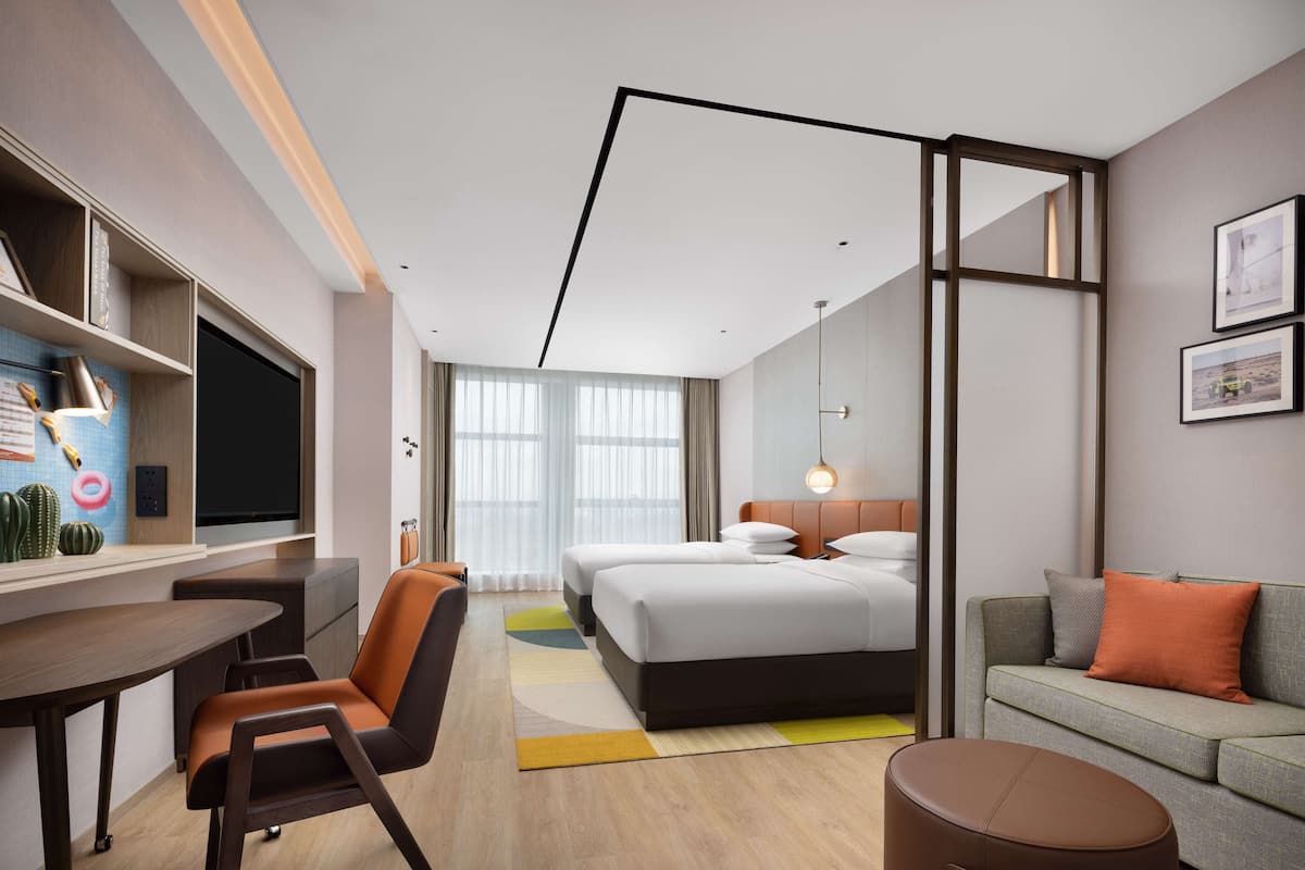 A modern hotel room with a bed, a sofa, a desk, and a chair. There is a window with curtains, a flat-screen TV, and a bookshelf with decorative items.