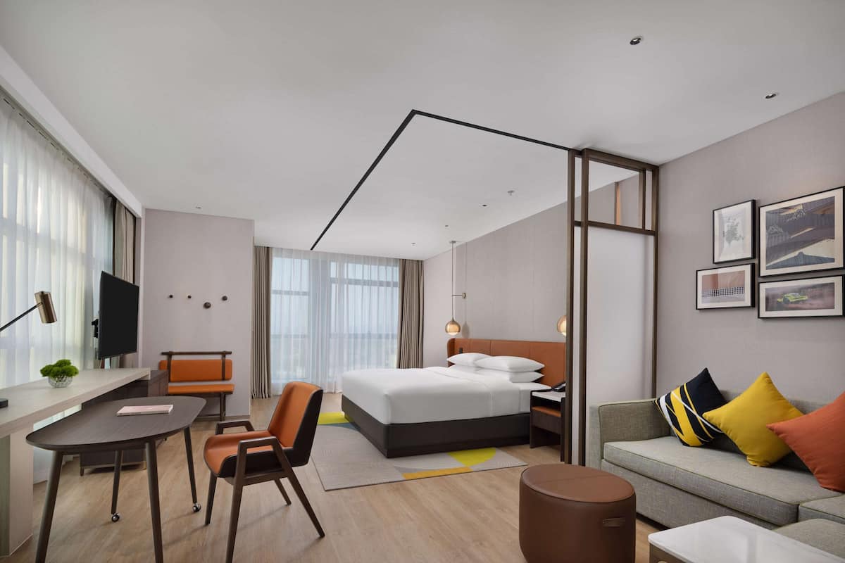 A modern hotel room with a bed, desk, chair, sofa, and a large window.