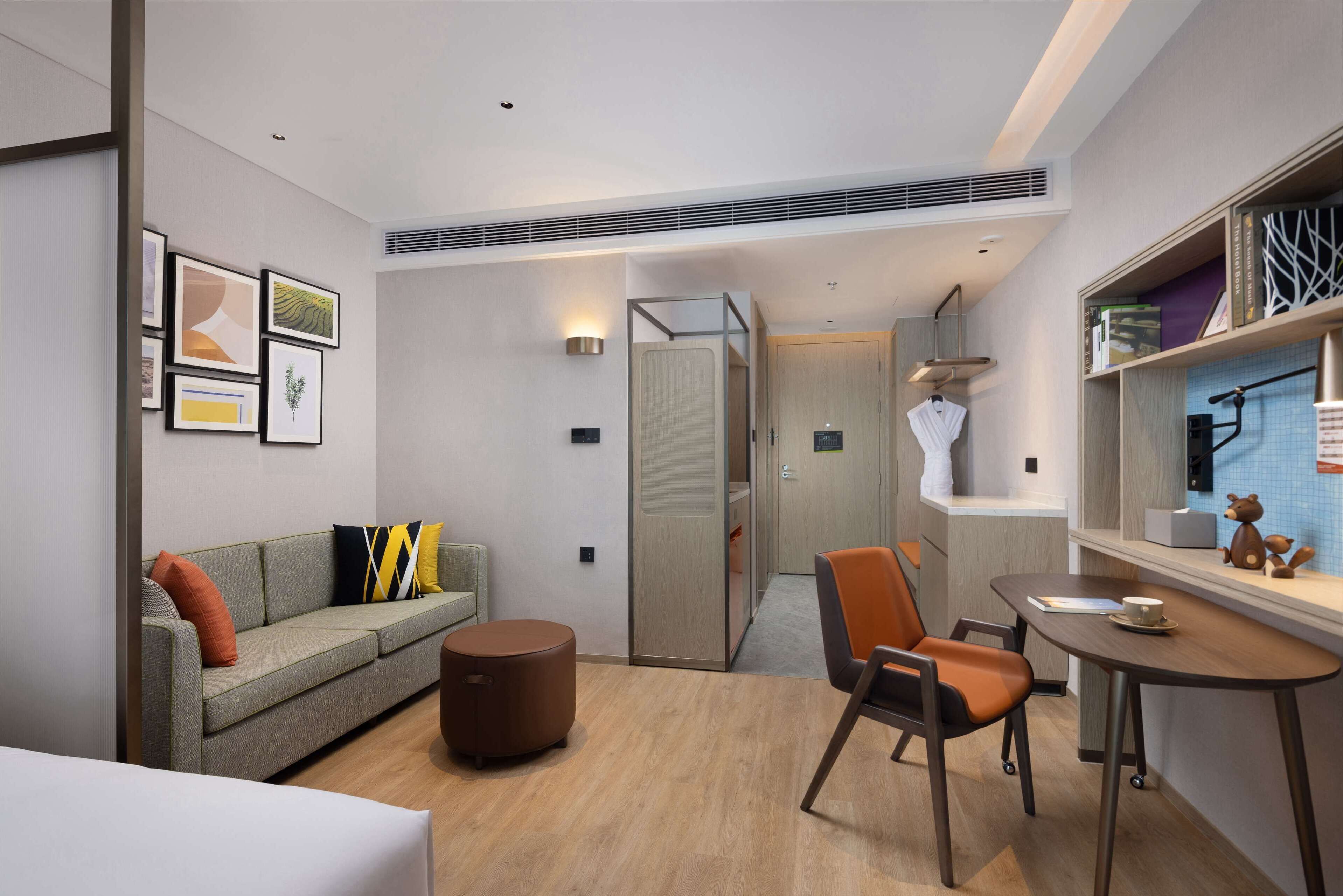 A modern hotel room with a sofa, a round ottoman, a dining table with chairs, and a small kitchen area.