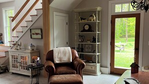 Een ruime woonkamer met een bruine leren fauteuil, een houten trap, een witte deur en uitzicht op een groen buitengebied.