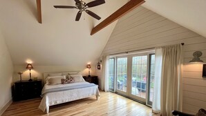 Een slaapkamer met een schuine plafond, houten vloer, een bed met wit beddengoed, twee bedlampjes en uitzicht door grote schuifdeuren op een terras.