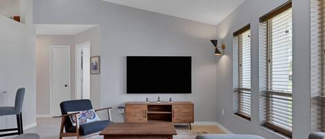 Een moderne woonkamer met een flatscreen televisie, een houten tv-meubel en een groot raam met jaloezieën.