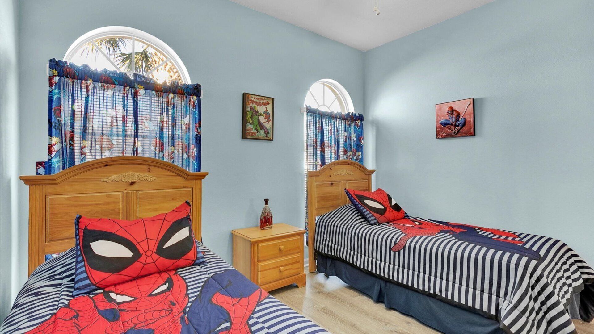 Une chambre avec deux lits, chacun recouvert d’une couette aux motifs de Spider-Man. Il y a deux tables de chevet, dont l’une porte une bouteille. Les murs de la chambre sont d’un bleu clair et il y a une fenêtre avec un rideau.