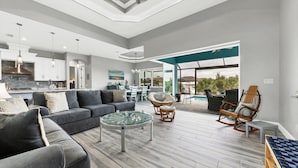A spacious living area with a grey sofa, a glass coffee table, wicker chairs, and a view of a swimming pool.