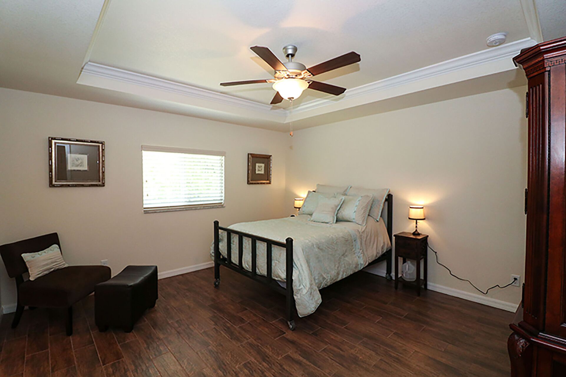 A bedroom with a bed, a chair, a small table, a ceiling fan, and a window with blinds.