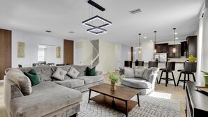 A modern living room with a sectional sofa, a coffee table, and a kitchen area in the background. - 5 Star Villa Serenade – A Symphony of Luxury (Orlando)