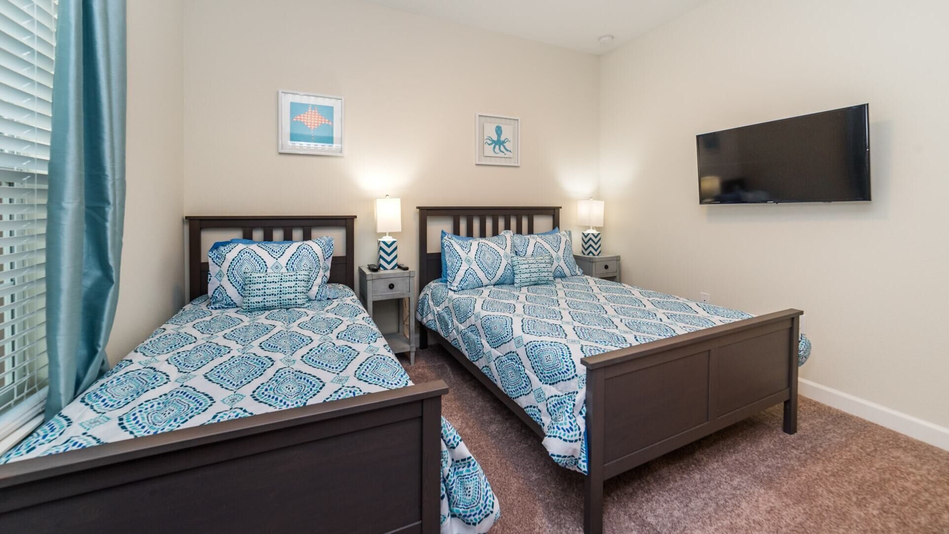 A hotel room with two beds, each with a patterned bedspread, bedside lamps, and a flat-screen TV mounted on the wall.