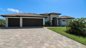 A modern two-story house with a brick driveway, garage doors, and a well-maintained lawn.